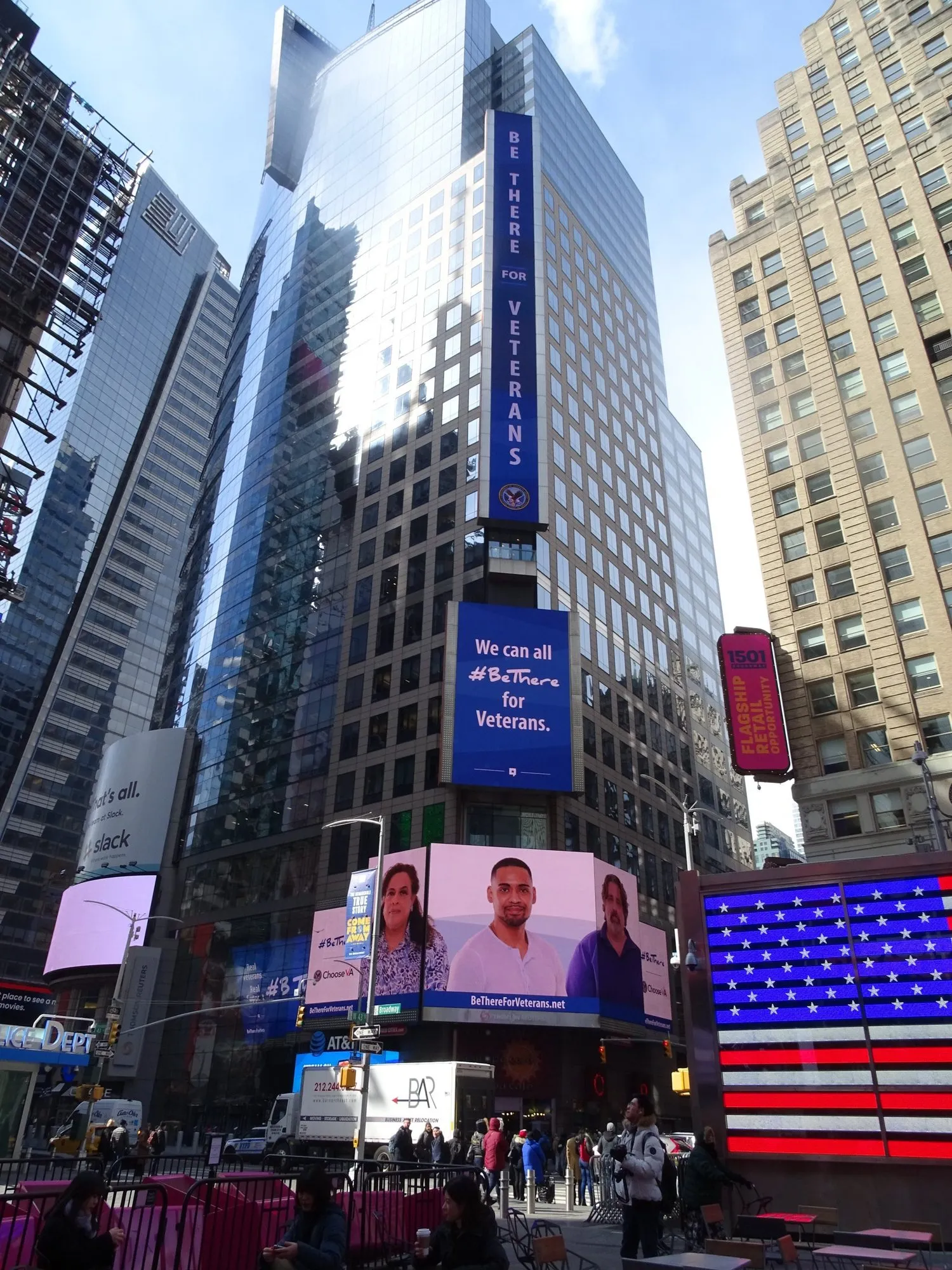 A vertical “Be There for Veterans” digital banner on a Times Square tower, a flag-lit façade below it.