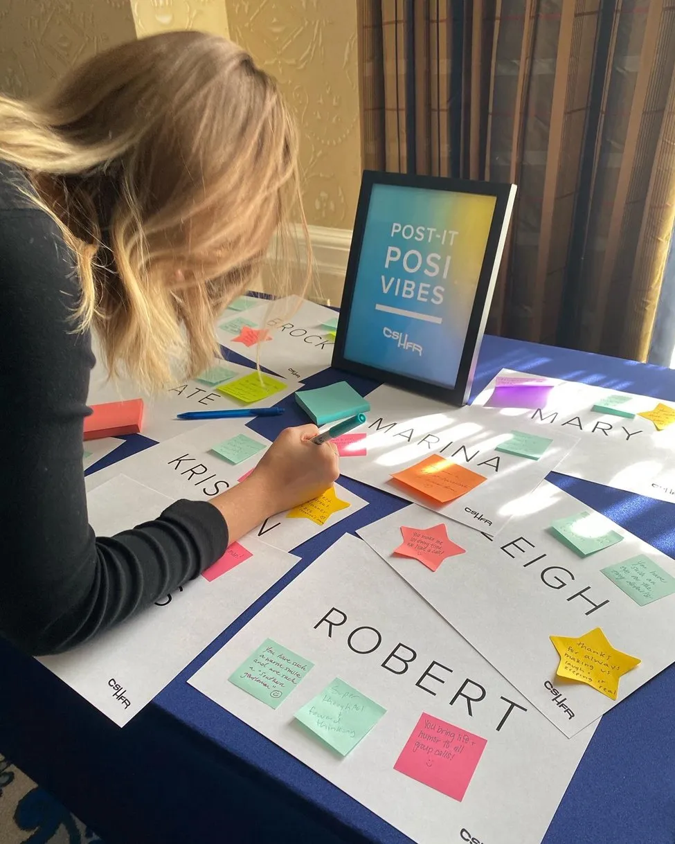 A retreat attendee adding sticky notes to a “Post-It Posi Vibes” board of hand-lettered name cards.